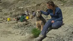 Bamforth ao lado da cadela Aster com pessoas escavando ao fundo