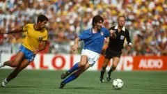 A imagem mostra um jogador da seleção italiana conduzindo a bola em velocidade durante uma partida contra o Brasil, enquanto é perseguido por um defensor brasileiro. Ao fundo, o árbitro acompanha o lance e as arquibancadas lotadas reforçam o clima de uma decisão em um grande estádio.
