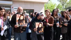 Kalabalık bir grup insan, önde iki kişi Rojvelat Kızmaz'ın fotoğrafını taşıyor. Bir kadın ağlıyor. Bir başka kadın elindeki mikrofonları ağlayan kadına doğru uzatıyor. 