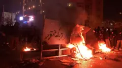 آتش روشن است و بر یک تابلوی شهری نوشته شده است مرگ بر دیکتاتور. جمعیت اطراف ایستاده‌اند و به نظر می‌رسد نرده‌های خیابان کنده شده است. 