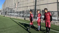 Três crianças palestinas de cerca de 10 anos, vestindo uniforme de futebol vermelho, aguardam na fila para bater um pênalti. Estão em um campo de grama sintética verde, com uma grade metálica atrás. Por trás da grade, avista-se um muro alto de concreto