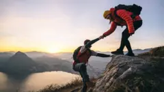 Mulher puxa homem para subir uma montanha em frente a um lago.