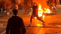 A masked protester raises his hand in font of a burning fire in the street at night