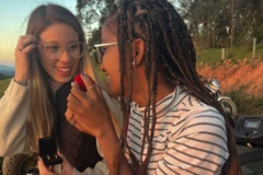 Fotografia colorida mostra duas mulheres sorrindo sentadas ao livre. Elas seguram caixinhas de anel de noivado e olham uma para outra