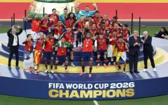 كابتن المنتخب الإسباني، رودري، يرفع كأس العالم، فيما يظهر خلفه لاعبو المنتخب الإسباني وهم يرتدون الميداليات الذهبية ويحتفلون بالفوز. كما يظهر في الصورة الرئيس الأمريكي، دونالد ترامب، ورئيس الاتحاد الدولي لكرة القدم، جياني إنفانتينو، عقب فوز إسبانيا في المباراة النهائية لكأس العالم 2026.