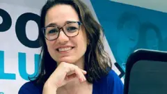 Mulher sorrindo para foto em escritório; ela está sentada, com a mão embaixo do queixo
