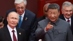 Perezida w'Uburusiya Vladimir Putin na Perezida w'Ubushinwa Xi Jinping bahagararanye. Xi atunze urutoki imbere, mu gihe Putin yerekeje amaso aho hantu yatunze urutoki, areba hejuru. Inyuma yabo hari abandi bagabo bahagaze. 