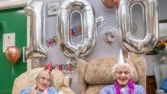 Duas senhoras sentadas sorrindo em frente a balões em formato de 100