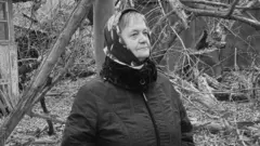 Чорно-білий портрет літньої жінки в куртці та хустці з квітами на тлі поламаних дерев закинутого подвірʼя