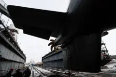 Большая подводная лодка в сухом доке, видна ее кормовая часть с большим гребным винтом и вертикальным рулем направления. Кругом металлические стены дока, рельсы для перемещения корабля, несколько человек в защитных касках. Освещение естественное, небо пасмурное.