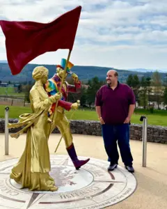 Daniel Marques de Araújo sorri ao lado de duas esculturas douradas que representam figuras em movimento, uma delas carregando uma grande bandeira vermelha. A cena acontece em um mirante ao ar livre, com colinas e áreas verdes ao fundo.