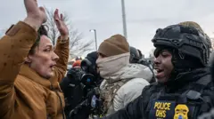 A demonstrator in a brown jacket holds their arms above their head in front of a uniformed official