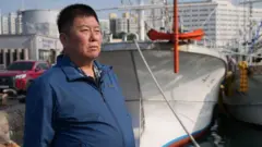 Um homem mais velho, de jaqueta azul, está em frente a um barco de pesca branco.