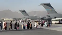 People carrying luggage walk across an airport tarmac towards large US military transport aircraft during an evacuation