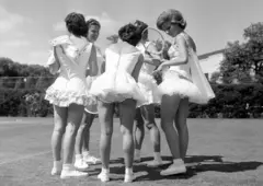 Fotografía en blanco y negro de cinco chicas conversando en círculo, todas con vestidos de tenis, mayoritariamente blancos y cortos, con volantes y encajes, mangas cortas. Todas calzan tenis y medias dobladas, sus piernas se ven casi completas. Se ven algunas raquetas. Están en una cancha de pasto y en el fondo se ve vegetación.