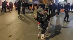 معترضان ایرانی در جریان یک اعتراض، با مسدود کردن خیابانی در کرمانشاه، ایران، در ۸ ژانویه ۲۰۲۶ تجمع کردند. در شب، معترضان خیابانی را مسدود کرده‌اند. در دوردست آتش‌های کوچکی دیده می‌شود و گروهی از مردم با لباس‌های تیره در خیابان تجمع کرده‌اند. در پیش‌زمینه، جوانی داخل یک چرخ‌دستی فروشگاهی نشسته و دستانش را بالا برده است؛ در حالی که مغازه‌های کرکره‌کشیده و نور چراغ‌های خیابان فضای پرتنشی ایجاد کرده‌اند.