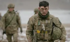 Karlı bir arka planda bir İngiliz komandosu kameraya bakarak yürüyor. Arkasında flu durumda başka askerler de var.