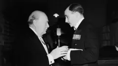 Dos hombres en perfil, vestidos de gala, conversan entre sí. El de la izquierda es más corpulento y tiene calvicie. El de la derecha es más alto, tiene bigote y condecoraciones en le pecho.