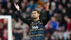 Iliman Ndiaye porte un maillot noir à rayures bleues et jaunes en zigzag. Il lève les bras en signe de célébration après avoir marqué un but.