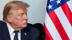 トランプ米大統領が黒い椅子に座って横を向いている写真。背後にはアメリカ国旗が置いてある