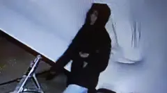 Imagem de uma câmera de segurança mostra uma pessoa com um casaco preto com capuz caminhando.