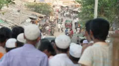 কক্সবাজারে রোহিঙ্গা ক্যাম্পের একটি রাস্তার ছবি, টুপি পরা কিছু মানুষ জড়ো হয়ে তাকিয়ে আছেন রাস্তার দিকে