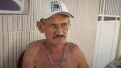 Homem de 72 anos, sem camisa, usando um cordão no pescoço e um boné branco