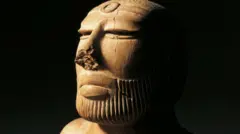 Uma estátua em close do rosto, com barba bem aparada, iluminada por luz amarelada contra um fundo preto. A escultura tem um círculo entalhado na testa, e o nariz parece parcialmente desgastado