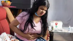 A mid adult Indian woman wearing surgical nose mask, sitting on sofa, feeling stomachache, health care concept