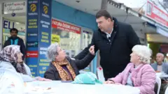 Yetişkin ayakta, üç ileri yaşlı kadın bir masada oturmuş, yüzleri ona dönük konuşuyorlar. 