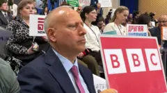 O correspondente da BBC Steve Rosenberg aparece em uma coletiva de imprensa segurando uma grande placa vermelha com a palavra “BBC”