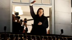 Siyah giymiş bir kadın otel balkonundan el sallıyor, arkada kaldığı odada fotoğrafçılar var