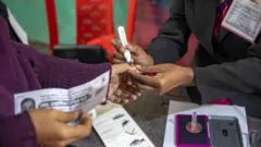 একজনের এক হাতে ব্যালট ও অন্য হাতের এক আঙুলে অমোচনীয় কালি দিচ্ছেন আরেকজন 