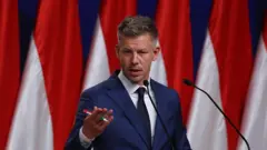 Молодий політик у темно-синьому костюмі стоїть за трибуною з мікрофонами та жестикулює рукою. У тлі — кілька вертикальних червоно-білих прапорів. 