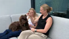 Kat Kitto sentada no sofá ao lado da filha Lily. Ela está dando comida para o cachorro