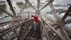 Jovem subindo as escadarias da Torre Eiffel, com vista panorâmica de Paris, na França