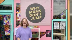 Helen Ledwick usa um suéter roxo e sorri diante de um pôster com os dizeres “Why Mums Don’t Jump” (Por que Mães Não Pulam, em tradução livre).