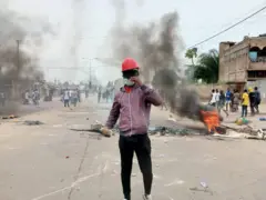 Un jeune manifestant avec casque rouge, visage caché et une pierre à la main droite. derrière lui des pneus brûlés avec des flammes, de la fumée noire et d'autres manifestants.