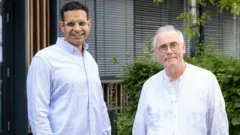 Khallaf, usando óculos e uma camisa listrada em azul e branco, e Lewin, de óculos e camisa branca, posam em frente a um edifício cercado por vegetação.