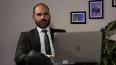 Eduardo Bolsonaro de terno e sentado à frente de um laptop, ao lado de uma planta e com um quadro da Estátua da Liberdade ao fundo