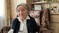 Wanda Traczyk-Stawska, de 98 anos, encara a câmera. Sentada em uma poltrona, veste uma blusa bordada e um cardigã