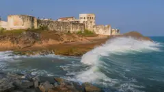 Vista lateral do exterior do Castelo de Cabo Corso com as ondas do Atlântico quebrando na praia abaixo das muralhas do edifício.
