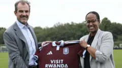 Umugabo n'umugore bafashe umupira wo kwambara utukura w'amaboko magufi wanditseho 'Visit Rwanda'. Barimo kumwenyura. Inyuma yabo hari ikibuga gitoshye.