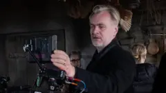 O diretor Christopher Nolan veste uma jaqueta preta e está no centro da imagem, olhando através de uma câmera IMAX