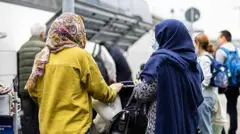 تصویر دو زن پناه‌جوی افغان در یکی از میدان‌های هوایی آلمان. این زنان به وسیله برنامه‌های پذیرش افغان‌ها به هانوفر آلمان منتقل شدند. پشت این زنان به سمت دوربین است. یکی شان لباس زرد روشن به تن دارد و دیگرش که مسن‌تر است لباس آبی با چادر آبی تیره به سر دارد. 