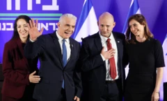 İki takım elbiseli adam ve eşleri sahnede, insanlara el sallıyor ve onlar selamlıyor.