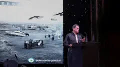 Abdi bir kürsüde konuşuyor arkasında duvara bir resim yansıtılmış, siyah beyaz bir şehri gösteriyor.