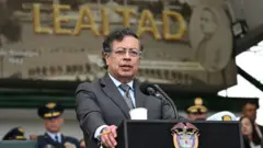 Gustavo Petro de pie en un podio mientras le habla a varios militares. Viste un saco gris y una corbata del mismo color. Su camisa es azul.