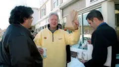 Band, vistiendo una chaqueta negra, está en la calle sosteniendo varios cafés para llevar mientras Clinton, vistiendo una sudadera amarilla, sostiene triunfalmente una bolsa de papel marrón y lleva un café.