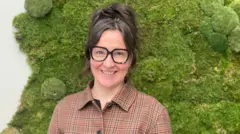 Jannie Jeppesen sorri para a câmera, usando uma camisa quadriculada e óculos pretos